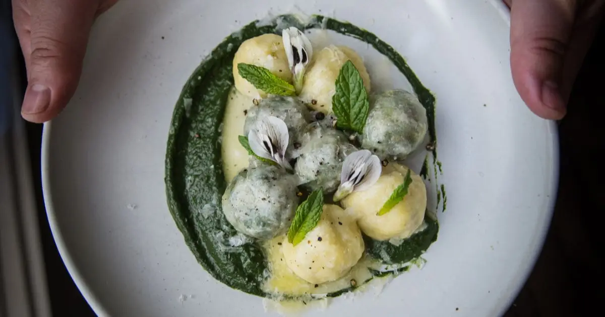 A white plate is held in two hands, presenting an artfully arranged dish of round, green and white gnocchi on a bed of green pesto. The dish, potentially the best dinner in San Francisco, is garnished with fresh mint leaves and delicate white flower petals.