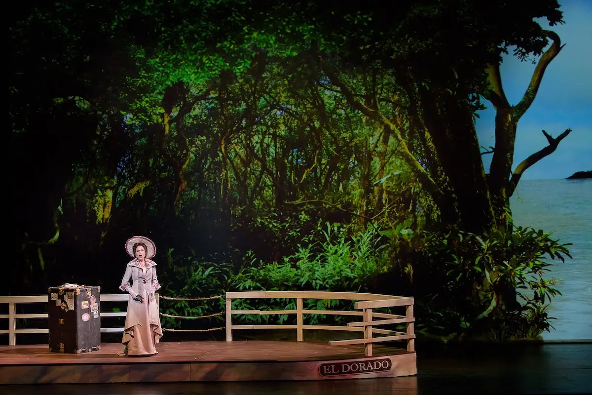 A person dressed in white, wearing a wide-brimmed hat, stands on a wooden stage marked "El Dorado" against a backdrop of a dense forest leading to the sea—one of the best things to do in South Bay this April. A steamer trunk sits beside them, completing the jungle-like scenery.