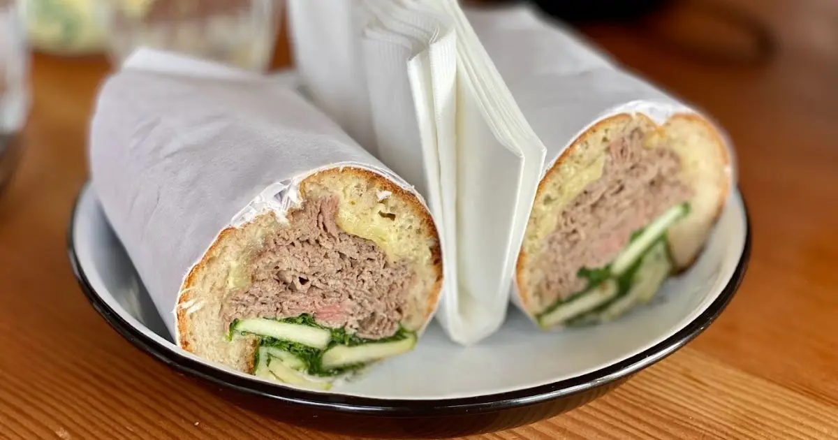 A sandwich cut in half is presented on a white plate with a black rim, making it the best lunch in San Francisco. Each half is wrapped in white parchment paper, exposing the fillings which include shredded meat, cucumber slices, and greens. A folded napkin is placed between the sandwich halves.