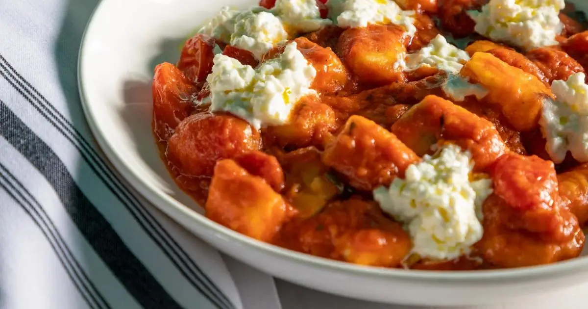 A white bowl is filled with golden-brown gnocchi coated in a rich, red tomato sauce. The dish, touted as the best dinner in San Francisco, is topped with dollops of creamy ricotta cheese. The bowl is placed on a white surface with part of a striped kitchen towel visible in the background.