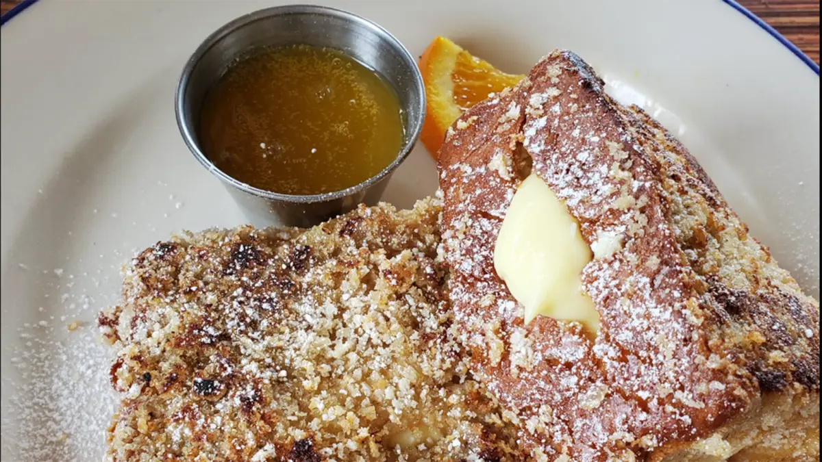 A close-up of a plate featuring French toast slices dusted with powdered sugar. One slice is topped with a pat of melting butter. To the side, a small metal cup holds a serving of syrup. A slice of orange garnishes the plate—truly the best breakfast Hilo has to offer.