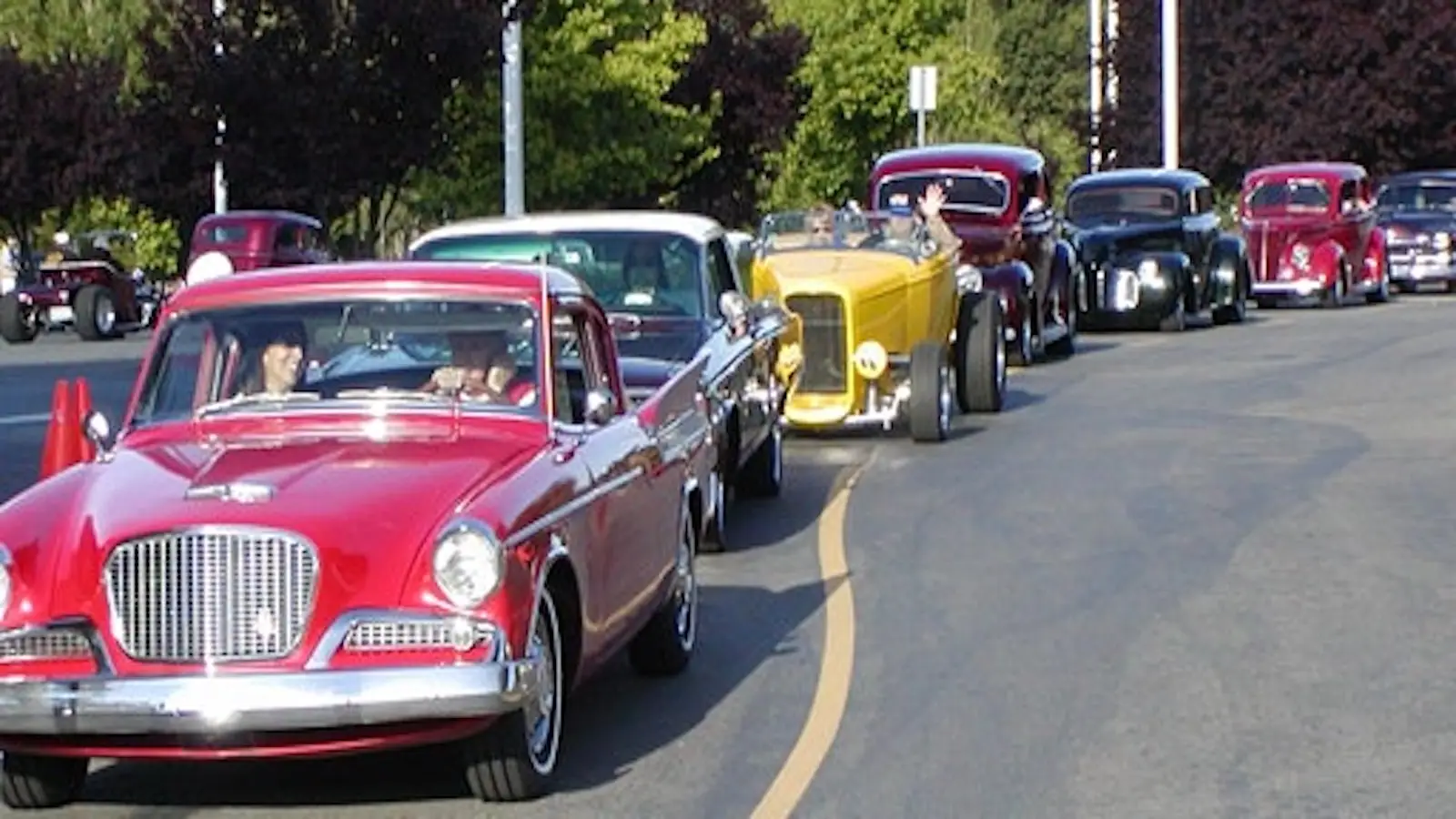 A line of classic cars drives down a curved road. The lead car is a red vintage vehicle followed by several other colorful classics, their passengers enjoying the ride. Trees line the background, evoking the charm of a car parade—a perfect June event and one of the best things to do in South Bay.