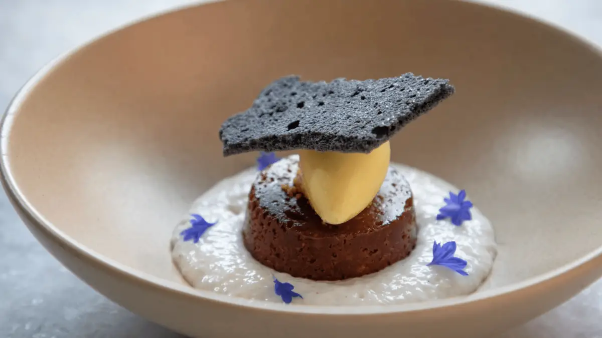 A dessert, touted as one of the Best Desserts in the Bay Area, is elegantly plated in a shallow beige bowl. It features a round cake topped with a quenelle of yellow ice cream or mousse, garnished with a black cracker. The cake sits on a bed of white creamy sauce, decorated with small blue edible flowers.