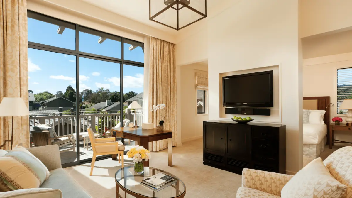 A well-lit living room with a modern design featuring a gray sofa, beige armchairs, and a round glass coffee table with a vase of flowers. A flat-screen TV is mounted on a black cabinet. Large windows with curtains open to views reminiscent of the Best Spa and Wellness Hotels in the Bay Area, overlooking trees and an inviting patio.