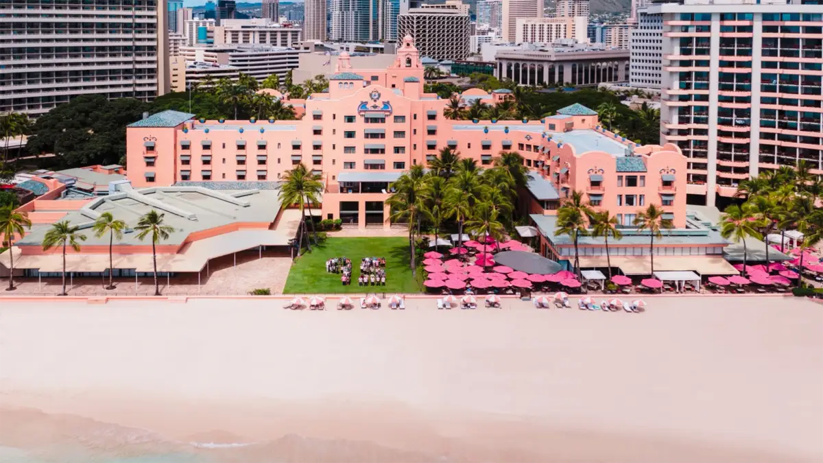 Aerial view of one of the best hotels on Oahu, a large pink beachfront hotel surrounded by palm trees and other buildings. The hotel has a green lawn with a group of people seated in the center. Beachfront umbrellas and lounge chairs are set up in front of the hotel on the sandy beach.