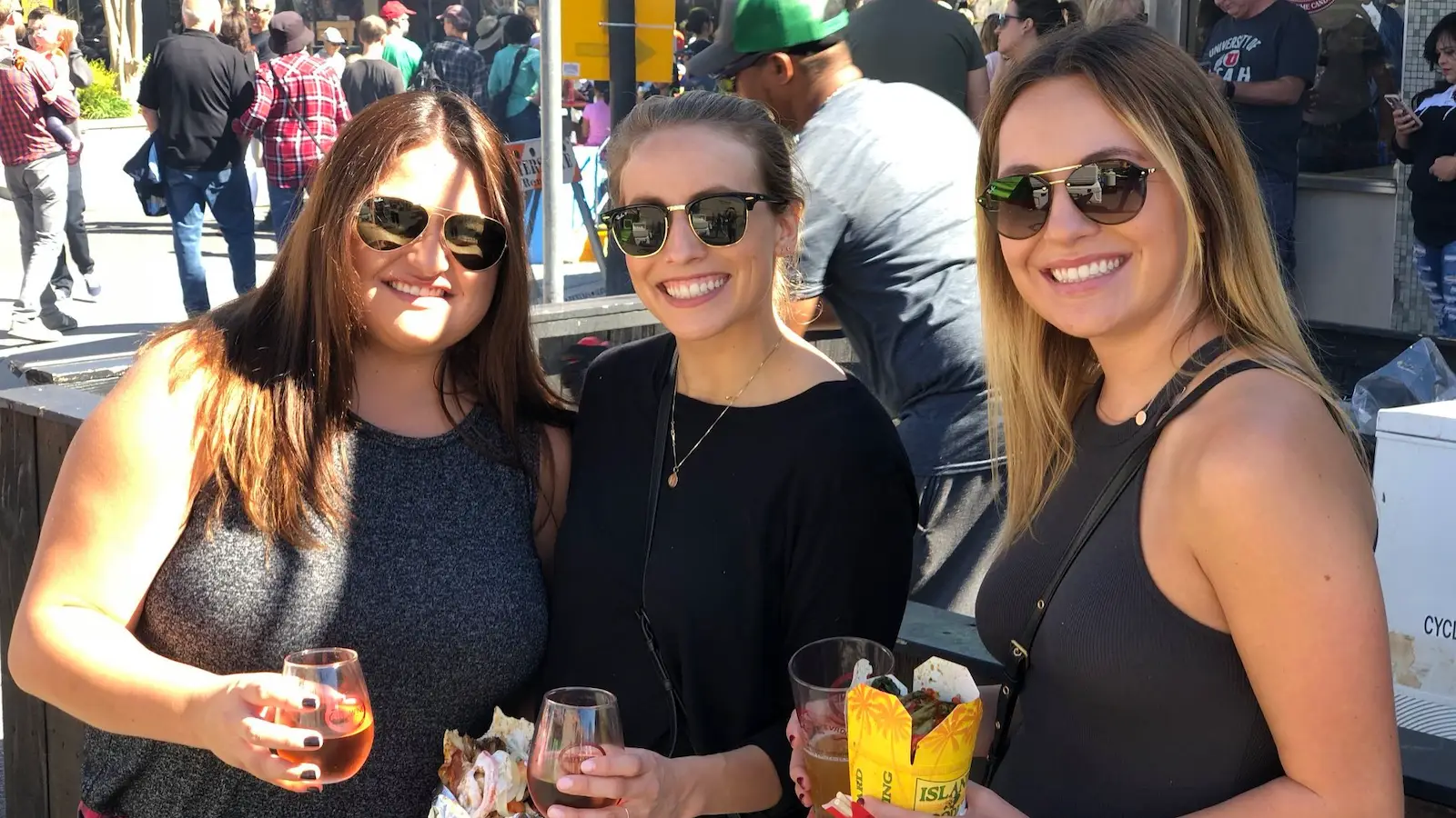 Three women are smiling and holding drinks at an outdoor event, one of the best things to do in South Bay this October. They are all wearing sunglasses and casual clothing, with a crowd of people and various event booths visible in the background. The atmosphere appears lively and sunny.