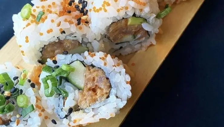 Close-up of the best sushi on the big island, beautifully presented on a wooden tray. The rolls, packed with avocado, cucumber, and a savory brown filling, are topped with sesame seeds, green onions, and vibrant fish roe. Seaweed is delicately wrapped inside the rice against a dark background.