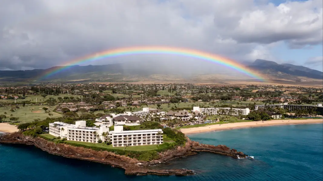 Aerial view of a coastal resort with several white buildings near a beach. A vibrant rainbow arches across the sky, framing green fields and mountains in the background. The ocean waters in the foreground are a deep blue, contrasting with the land—a scene akin to the best Oceanfront Hotels Maui West has to offer.