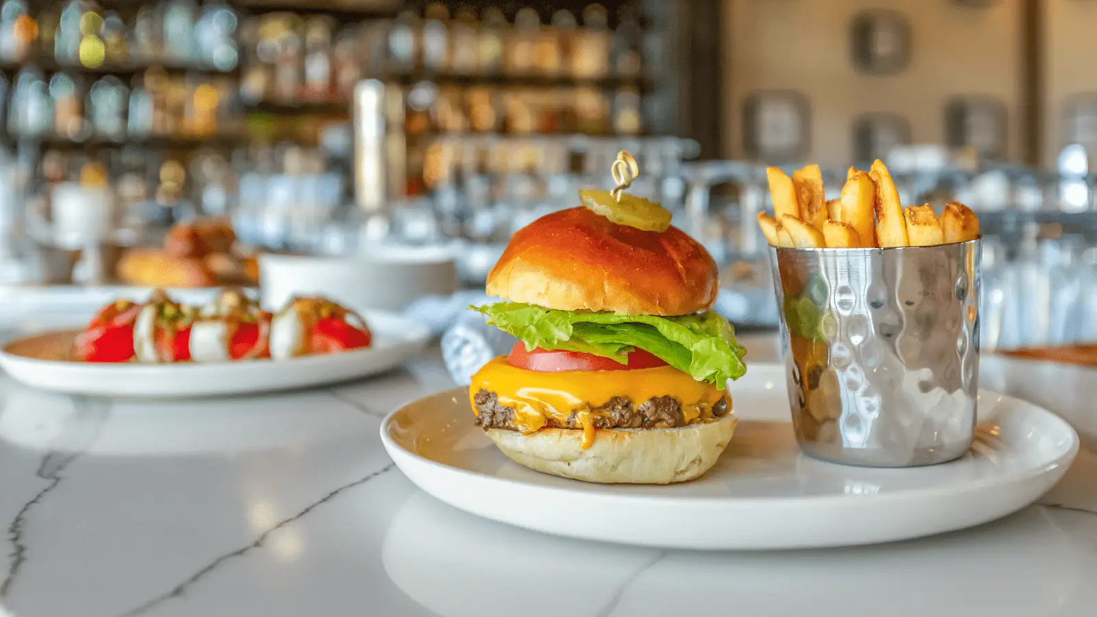 A cheeseburger with a lettuce leaf, tomato slice, cheese, and pickles on a bun is served on a white plate next to a small metal cup filled with French fries. In the background, out-of-focus plates of food and a bar shelf with bottles are visible—one of the best things to do in Sonoma County this February.