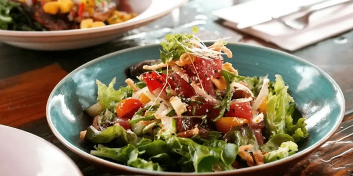 A colorful, fresh salad in a turquoise bowl featuring mixed greens, cherry tomatoes, cucumber, and slices of seared tuna, topped with sprouts and sesame seeds. In the background, another dish and cutlery hint at the best dinner on the North Shore waiting to be enjoyed on a wooden table.