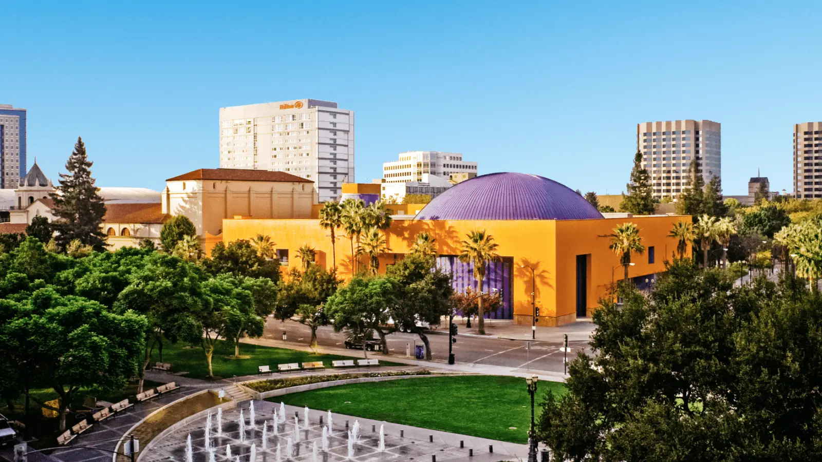 A vibrant Bay Area cityscape featuring a yellow and purple domed structure surrounded by lush green trees. High-rise buildings are visible in the background under a clear blue sky. In the foreground, a plaza with benches and a circular fountain can be seen.