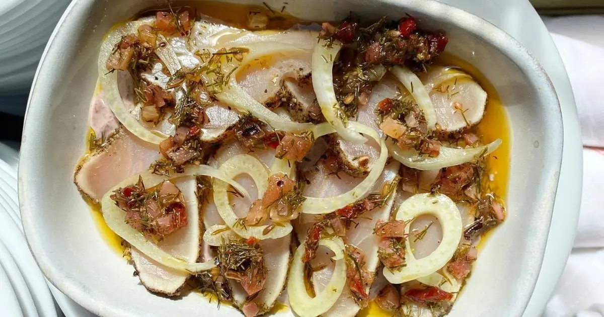 A white bowl containing a dish of thinly sliced raw fish topped with sliced white onions and a mix of herbs and diced vegetables in a light sauce. The fresh, colorful dish, garnished with finely chopped greens and red peppers, is among the best dinner options in San Francisco.