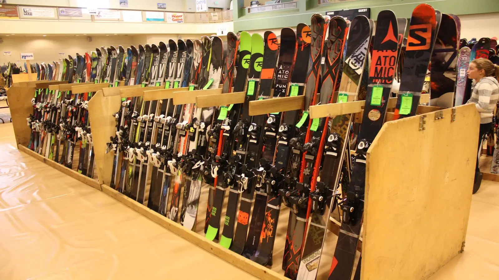 A selection of various skis displayed vertically in wooden racks at an indoor venue. Each pair of skis has bindings attached and green price tags. A person stands to the right of the racks, browsing through the selection, highlighting one of the best things to do in Tahoe November.