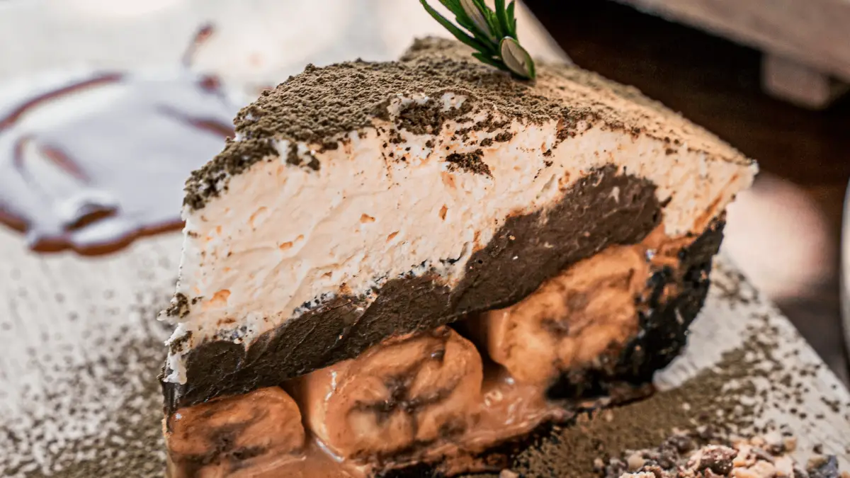 A slice of dessert featuring layers of whipped cream, chocolate filling, and visible banana slices on the bottom. The dessert is dusted with a green powdered topping and garnished with a sprig of herbs on a plate with a caramel drizzle. Truly one of the Best Desserts in the Bay Area!