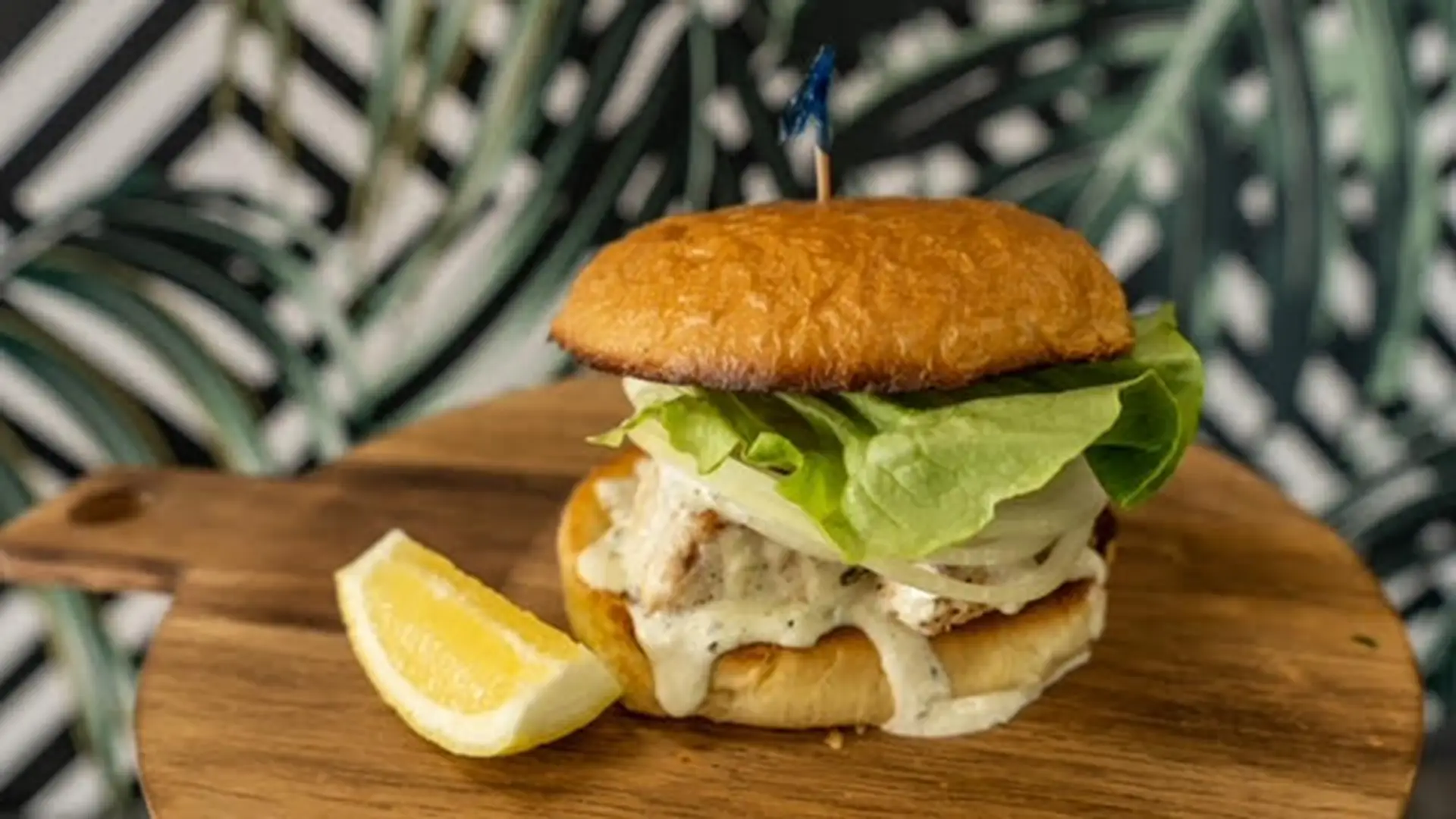 A fish sandwich with a golden-brown bun, lettuce, creamy white sauce, and a thick fish fillet, secured with a toothpick on a wooden board with a lemon wedge. The background features a patterned leafy design. This culinary delight could easily compete for the best burger on the big island.