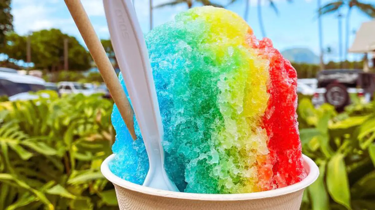 A vibrant, rainbow-colored shaved ice dessert in a white paper cup, placed outdoors, showcasing the best Hawaiian food. The ice is shaded with reds, yellows, greens, and blues and comes with a white plastic spoon and a wooden stick. Lush greenery and palm trees are visible in the background.