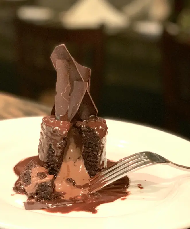 A decadent chocolate dessert, one of the best desserts in the Bay Area, sits on a white plate. The dessert features a rich chocolate cake with a molten chocolate center, topped with shards of chocolate. A fork is partially slicing through the cake, revealing its gooey interior.