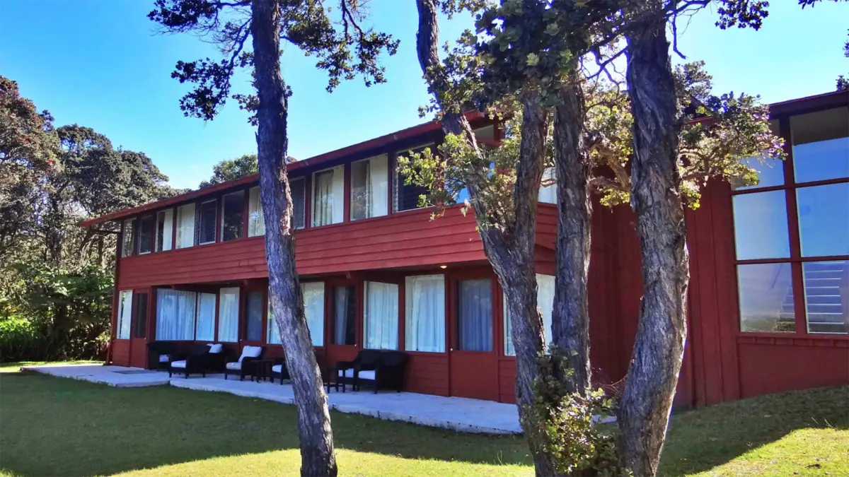 A two-story red wooden volcano house with large windows and a row of black and white outdoor chairs on the ground floor porch. The house is surrounded by a green lawn with several slender trees in the foreground. It's a sunny day with a clear blue sky.