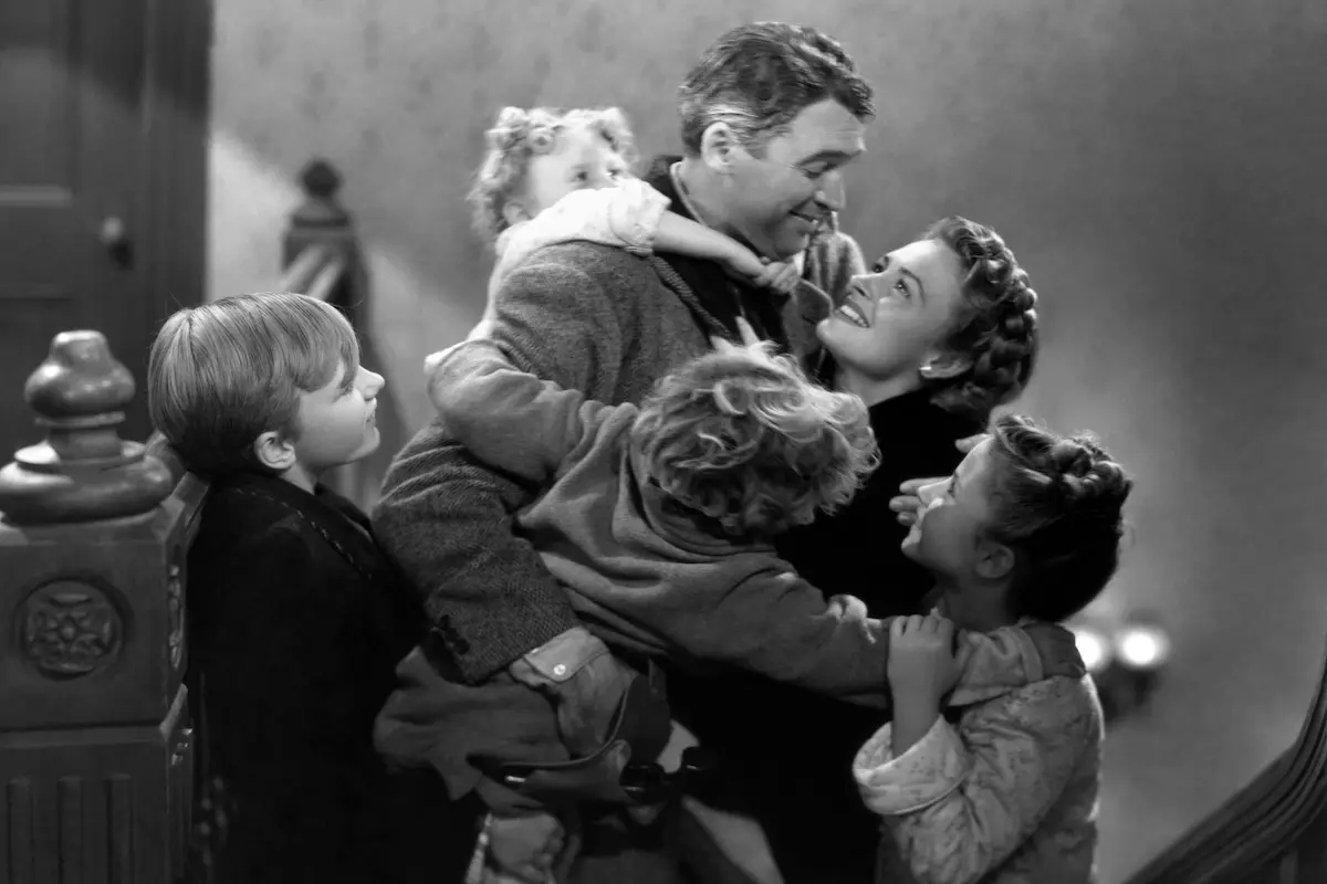 A black-and-white image depicts a joyful family embracing on a staircase. A man and woman, dressed in mid-20th-century clothes, are surrounded by four children who appear delighted and affectionate, clinging to their parents with smiles and laughter—perfect for The Ultimate Holiday Movie List.