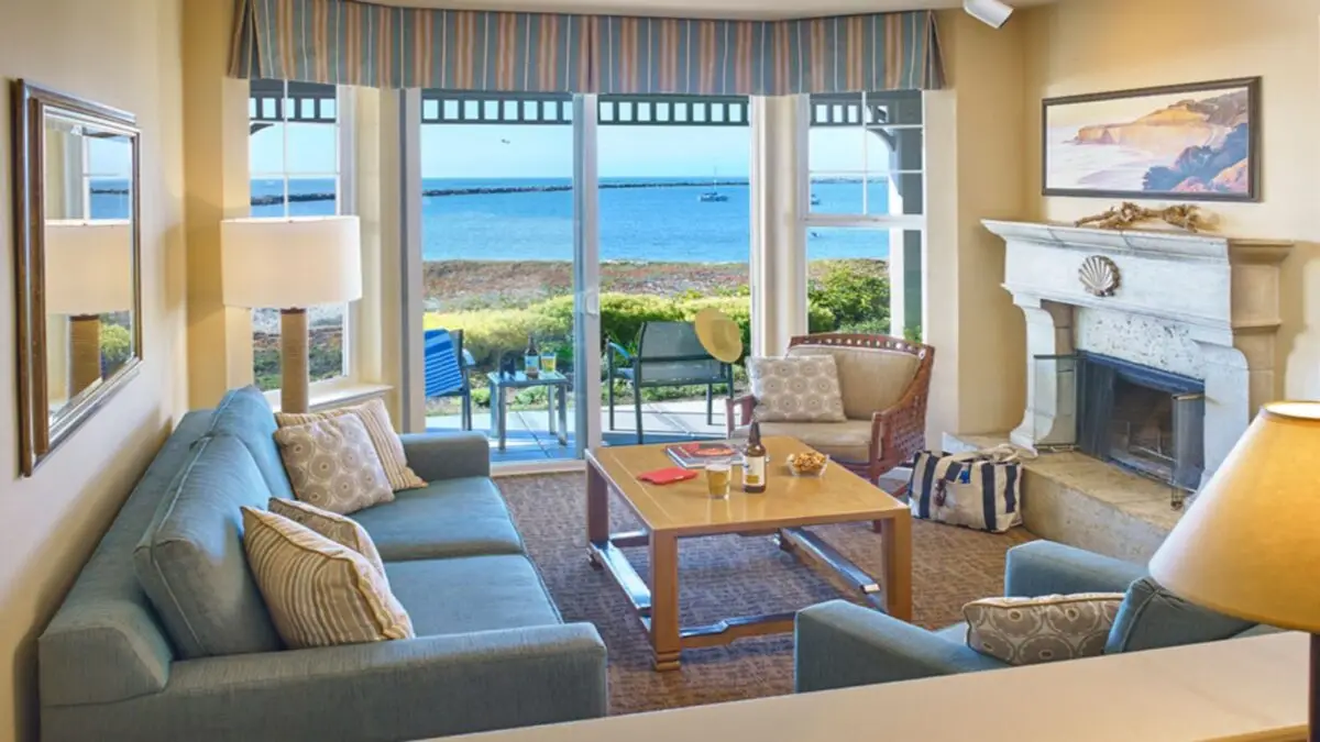 A cozy beach house living room in Half Moon Bay featuring a blue sofa, armchairs, a wooden coffee table, and a fireplace with a mantle. Large windows offer a scenic view of the ocean and rocky shoreline. A small table with chairs is set up outside on the patio. The room is warmly lit.