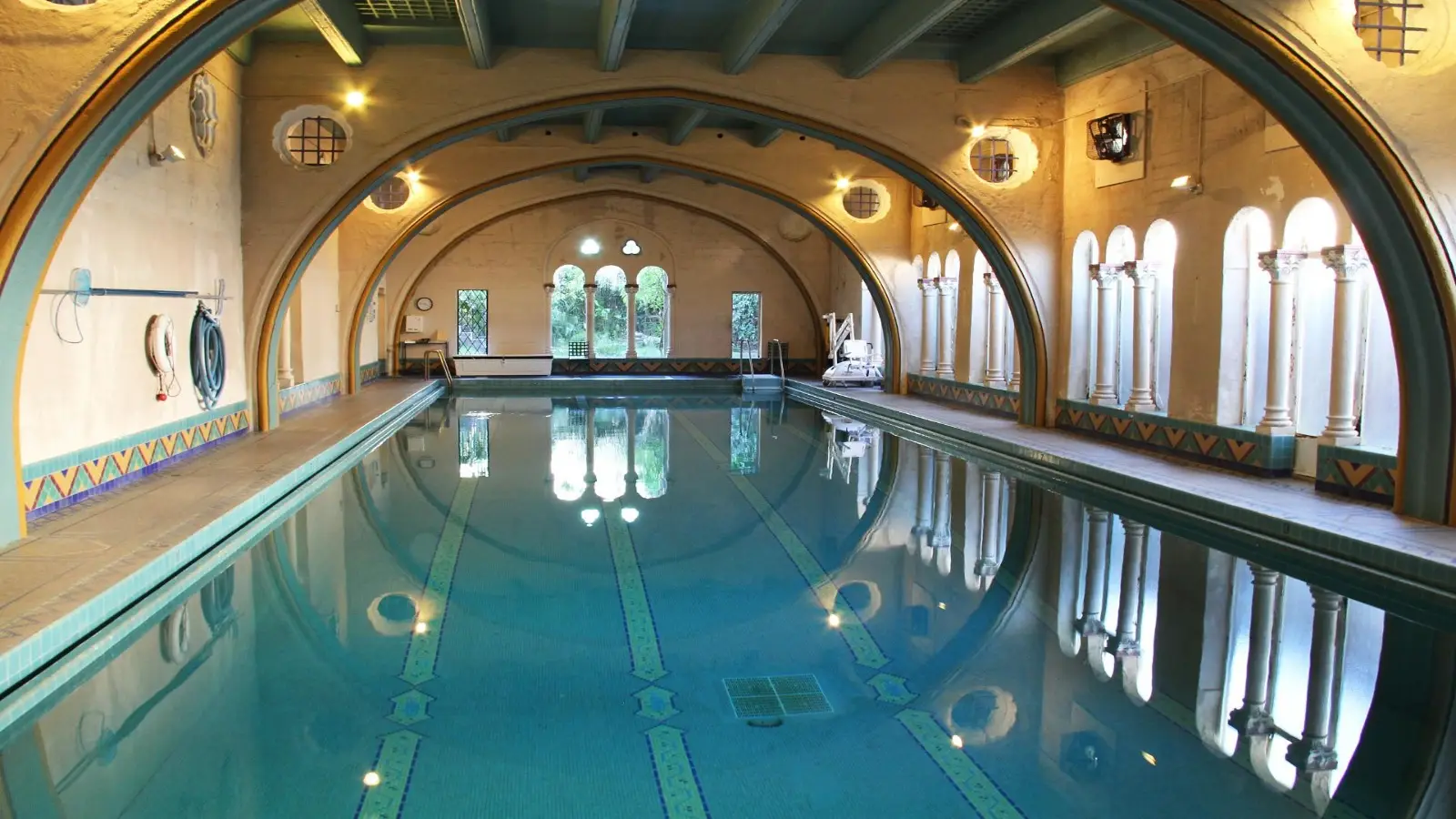 A luxurious indoor pool with arched ceilings, ornate columns, and large windows letting in natural light awaits you at one of the finest local getaways. The pool has two swimming lanes and intricate tile patterns. The elegant room features calming, ambient lighting that enhances the serene atmosphere.