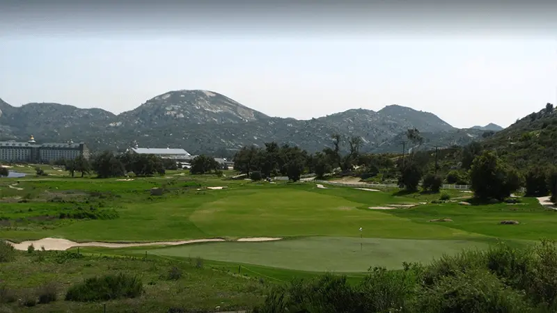 A scenic view of one of the best golf courses in the Bay Area, featuring well-maintained green grass, situated in a valley surrounded by rugged, rocky mountains. Trees and shrubs are scattered around the course, and a white building is visible in the distance under a clear sky.