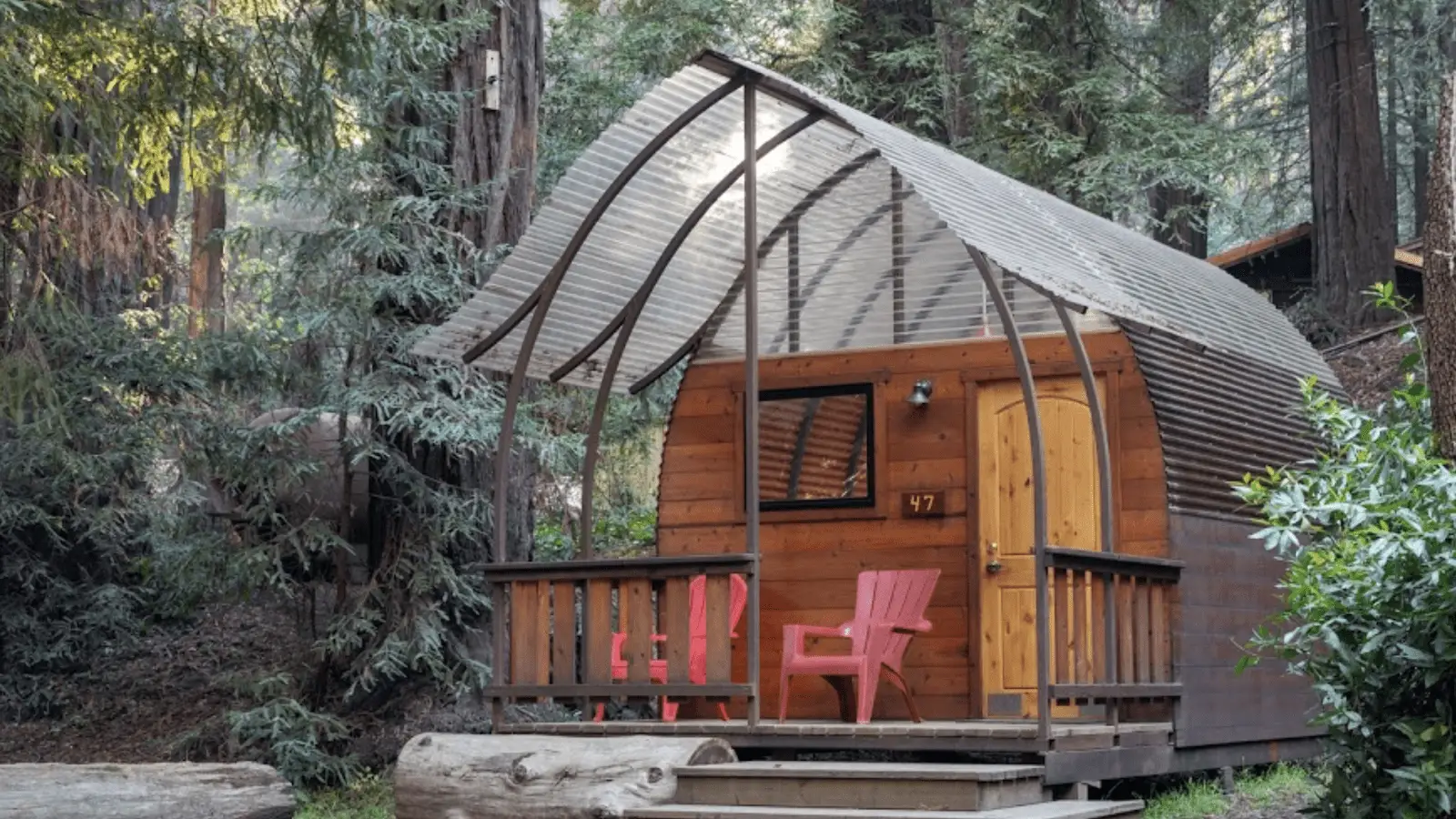 A small, rustic wooden cabin with a curved metal roof stands among tall trees on the Monterey Peninsula. Two red Adirondack chairs sit on the front porch alongside a small wooden table, creating a cozy outdoor seating area. The cabin is surrounded by lush greenery and forest.