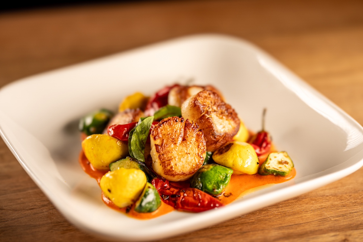 A gourmet dish featuring seared scallops, accompanied by a colorful assortment of yellow squash, Brussels sprouts, and cherry tomatoes. All ingredients are garnished with a light sauce and elegantly presented on a white square plate—perfect for the Mill Valley Restaurant Rally or any Bay Area brunch.