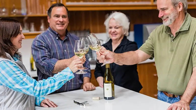 Four people smiling and toasting with glasses of white wine around a table. A bottle of wine and a corkscrew are on the table. The casual setting, with a wooden background and shelves in the backdrop, perfectly captures one of the best things to do in wine country. The group looks cheerful and relaxed.