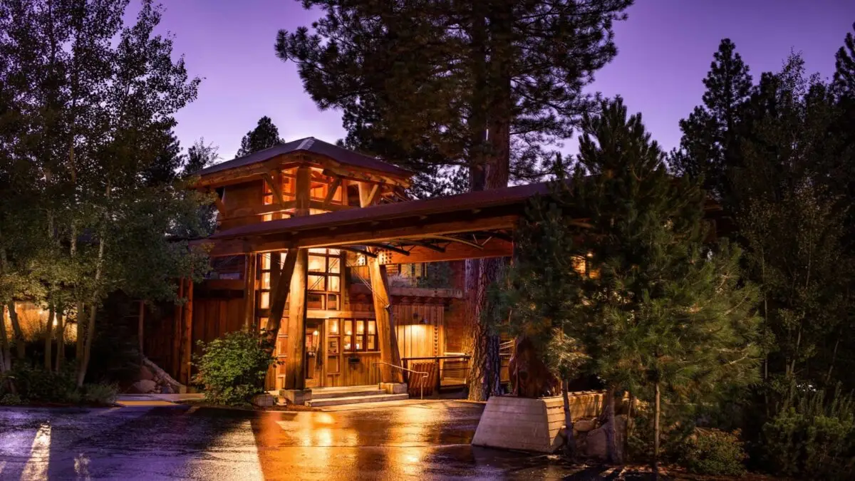 A cozy, wooden lodge nestled among tall pine trees glows warmly with yellow lights at dusk. The wet ground reflects the lights, enhancing the serene ambiance. The sky is tinged with a soft purple hue, adding to the tranquil and inviting atmosphere, making it one of the best hotels Tahoe offers.