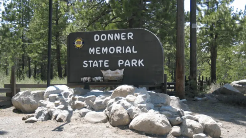 A wooden sign for Donner Memorial State Park stands amidst a pile of large rocks. The sign displays a small covered wagon at the bottom and is surrounded by tall green pine trees, with a sunny, clear sky in the background. It's one of the best places to visit near Lake Tahoe for outdoor activities and exploration.