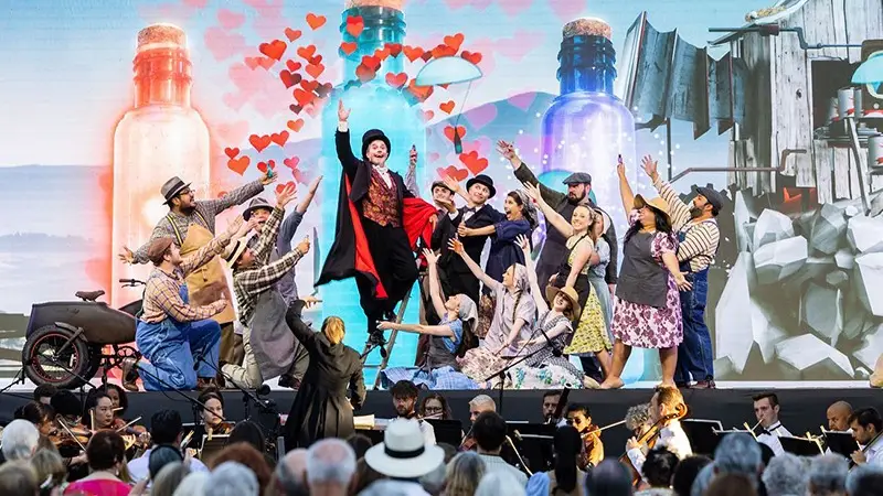 A vibrant stage performance, one of the best things to do in Wine Country this July, features a lively group of actors in colorful costumes, with one central figure in a red and black outfit. The backdrop shows large potion bottles with floating hearts. An orchestra plays in the foreground, while a captivated audience watches.