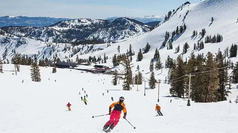 A skier in an orange and black jacket, and pink pants, descends a snowy slope. Numerous other skiers and snowboarders are scattered below. Snow-covered mountains and trees can be seen under a clear blue sky, showcasing one of the Best Places to Visit—Lake Tahoe—for outdoor activities.