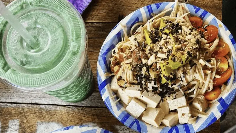 A bowl filled with a mix of noodles, tofu cubes, cherry tomatoes, sliced avocado, and sprinkled with chopped nuts and seeds is placed on a wooden table. A green smoothie in a plastic cup with a straw is set beside the bowl, reminiscent of dishes you'd find at the top vegan restaurants in Oahu.