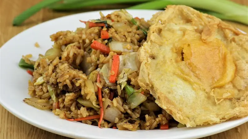 A plate of fried rice mixed with vegetables, such as carrots and leafy greens, is garnished with a crispy fried egg on the side. The dish, reminiscent of what you might find at top vegan restaurants in Oahu, is served on a white plate placed on a wooden surface, with green onions visible in the background.
