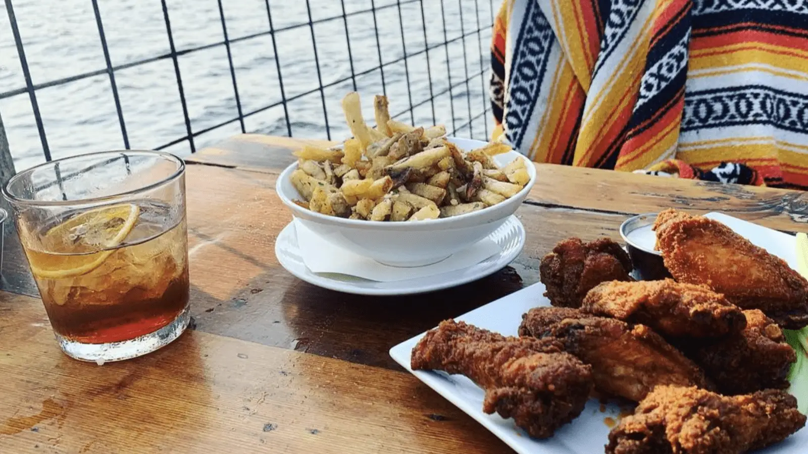 A close-up of a meal on a wooden table. There is a glass of dark liquid with a lemon slice, a bowl of seasoned fries, and a plate of crispy fried chicken. The background features a person wearing a colorful striped blanket and a view of water beyond a wire fence.