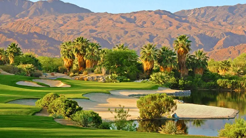 A pristine golf course, regarded as one of the best golf courses Palm Springs has to offer, features well-manicured greens, sand traps, and a reflective water hazard surrounded by lush palm trees and desert vegetation. In the background, rugged brown mountains rise under a clear blue sky. Golfing here is one of the top things to do in Palm Springs.