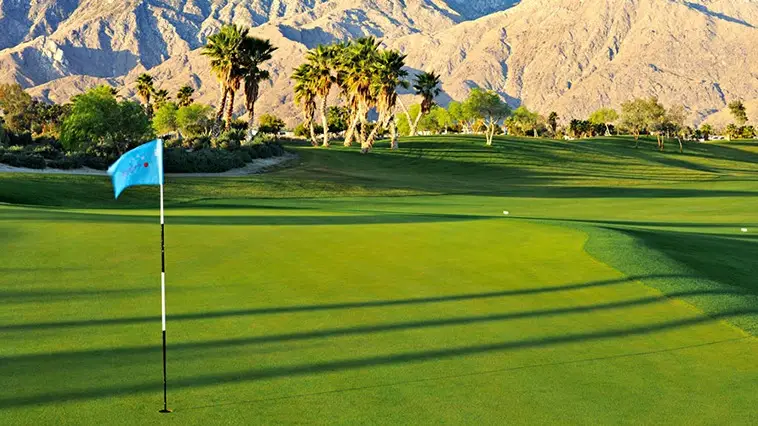 A golf course with lush green grass, a flag on the green, and a scenic backdrop of mountains and palm trees bathed in sunlight. The long shadows cast by the setting sun add depth to the landscape. Experience one of the best golf courses Palm Springs has to offer—the perfect addition to your list of things to do in Palm Springs.