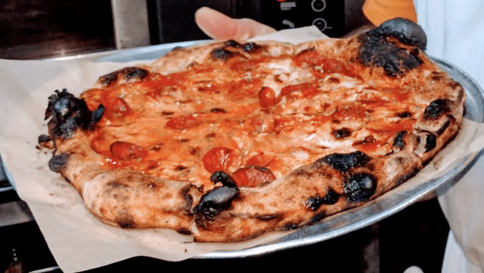 A person holding a freshly baked, slightly charred pizza topped with tomatoes and melted cheese on a metal tray lined with parchment paper. The pizza has a rustic, uneven appearance and a crispy, browned crust.