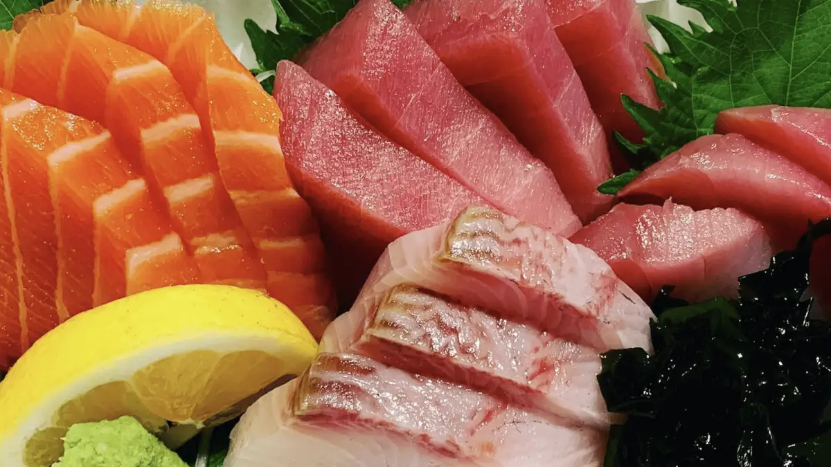 A plate of sashimi from the best sushi in North Bay features neatly arranged slices of raw salmon, tuna, and white fish. The fish slices are vibrant and fresh, accompanied by a garnish of green leaves, a lemon wedge, wasabi, and a side of seaweed.