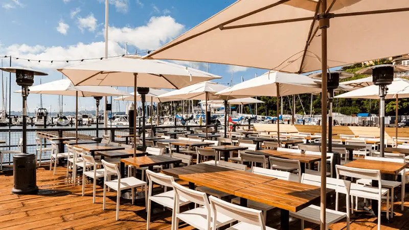 Outdoor dining area with wooden tables and white chairs under large beige umbrellas. The area overlooks a marina with numerous docked boats. Strings of lights hang above the seating area, and the sky is clear with few clouds.