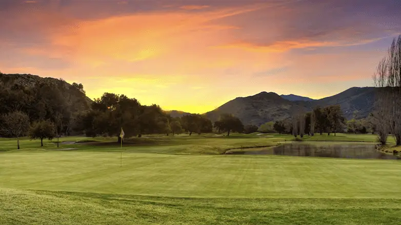 A scenic view of a golf course at sunset. The sky is painted with vibrant hues of orange, pink, and purple. The green fairway and the surrounding trees are bathed in soft light. A small pond reflects the colors of the sky, making it one of the best golf courses Bay Area offers.