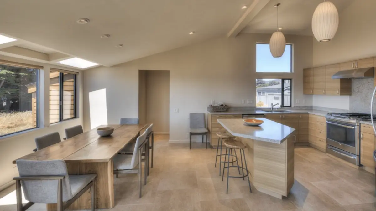 A modern, sunlit kitchen and dining area with beige tile flooring. The kitchen has light wood cabinets, stainless steel appliances, and a granite island with bar stools. The dining table is set with gray upholstered chairs. Pendant lights hang from the ceiling—reminiscent of the tasteful elegance found in the Best Group Getaway Hotels Sonoma.