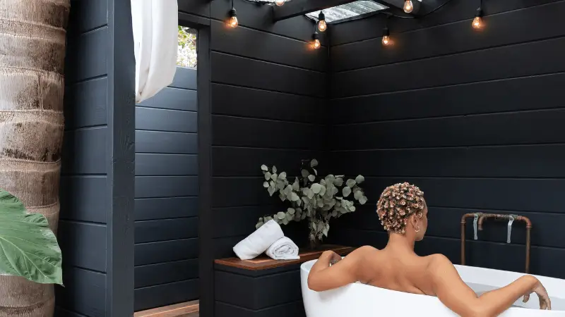 A person with short curly hair relaxes in a white bathtub in an outdoor setting with dark wooden panels, perfect for local getaways. There's a large plant in the foreground, a potted plant in the background, rolled towels, and string lights hanging overhead.