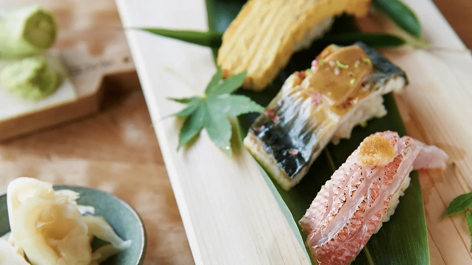 A close-up of elegant sushi pieces on a wooden platter garnished with green leaves. The best sushi in North Bay includes a piece of fish with a golden topping, a piece of thermidor grilled with egg, and a dish of pickled ginger and wasabi in the background.