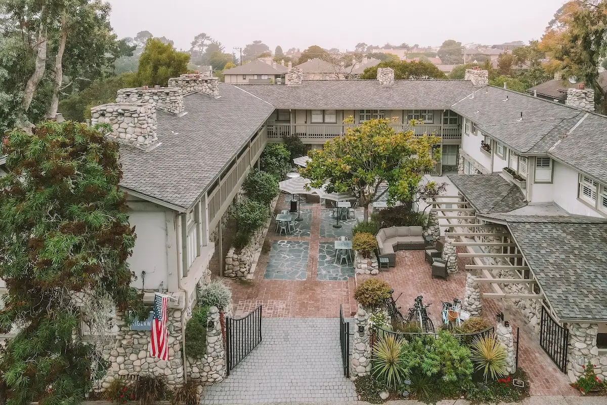 Aerial view of a rustic courtyard on the Monterey Peninsula, surrounded by stone and wood buildings with grey roofs. The courtyard features outdoor seating with tables and chairs, lush greenery, and a flagpole flying the American flag. Trees and other buildings are visible in the background.