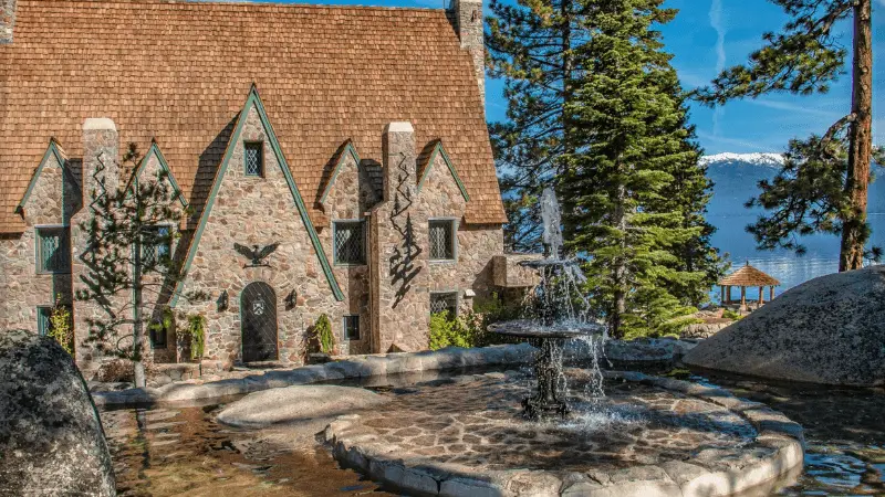 A picturesque stone cottage with a steep, shingled roof is surrounded by tall trees. In the foreground, a circular fountain with a statue of an eagle sprays water into a stone basin. A lake and snow-capped mountains are visible in the background—the perfect retreat to explore Tahoe's best places to visit.