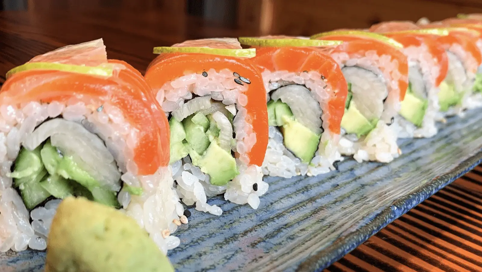 Close-up of a line of sushi rolls on a blue ceramic platter. Each roll has a filling of avocado and cucumber, topped with a slice of salmon, and a thin lemon slice. Wasabi paste is visible on the side. The table beneath the platter is wooden—truly the best sushi North Bay has to offer.