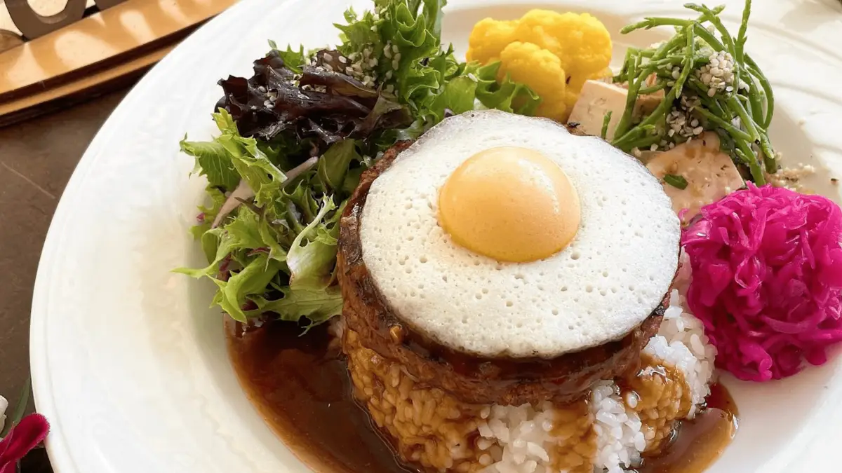 A gourmet dish features a piece of meat topped with a sunny-side-up egg, resting on a bed of rice with sauce. This culinary masterpiece, inspired by foods that define Hawaiian culture, is garnished with greens, yellow cauliflower, tofu pieces, and a bright pink flower, all carefully arranged on a white plate.