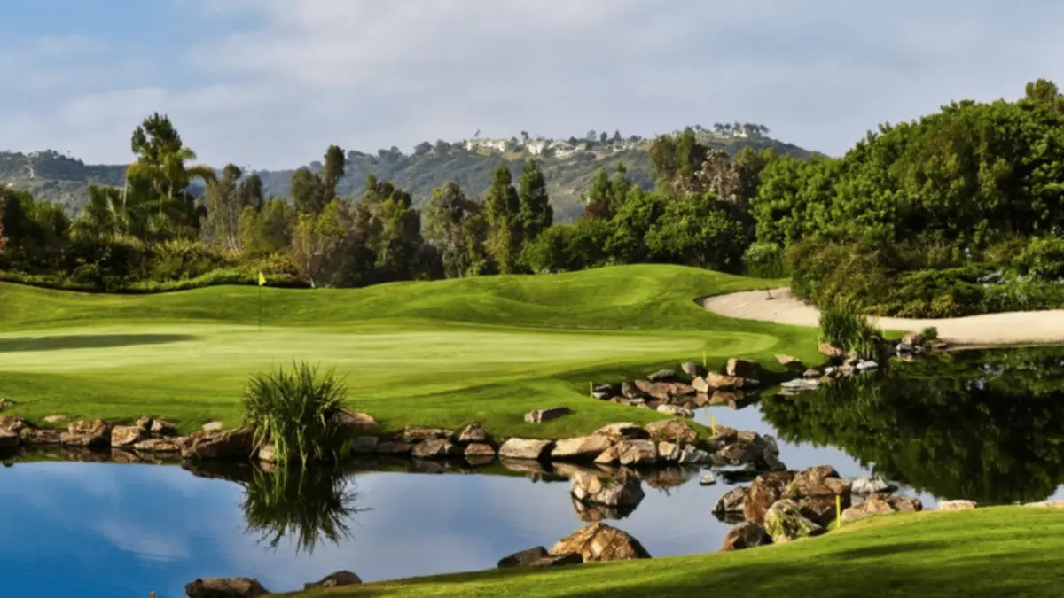 A scenic view of a golf course with a well-manicured green surrounded by lush trees and bushes. A small pond with rocks borders the green, reflecting the surroundings. In the background, rolling hills and a few scattered buildings are visible under a clear sky, making it one of the best golf courses Bay Area has to offer.